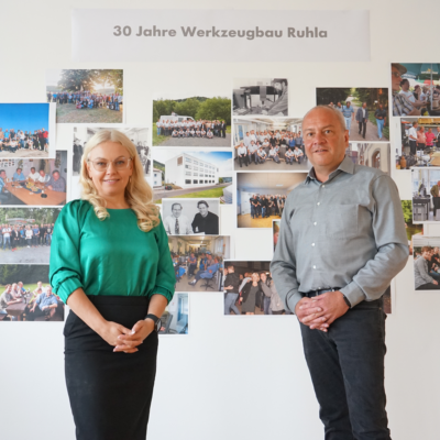 Die Geschäftsführer der Werkzeugbau Ruhla GmbH, Lena Lüneburger und Udo Köllner, vor der Fotowand des Unternehmens zum diesjährigen 30-jährigen Firmenjubiläum.