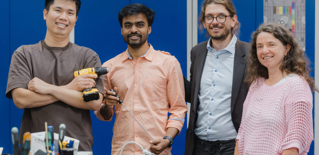 Fünf Mitarbeitende stehen gemeinsam in einer Werkstatt, zwei von ihnen halten Werkzeuge in der Hand, vor ihnen liegen elektronische Bauteile und Arbeitsmaterialien.