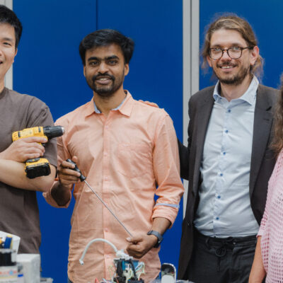Fünf Mitarbeitende stehen gemeinsam in einer Werkstatt, zwei von ihnen halten Werkzeuge in der Hand, vor ihnen liegen elektronische Bauteile und Arbeitsmaterialien.