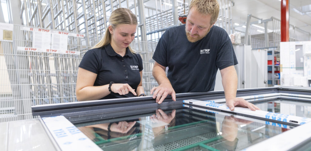 Eine Frau und ein Mann stehen nebeneinander in einer Produktionshalle. Vor ihnen liegt ein Fenster.