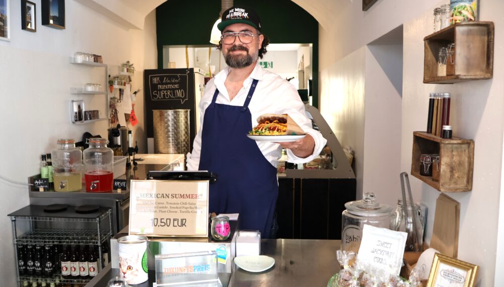 Ein Mann steht hinter der Theke und hält einen Teller mit einem Sandwich in der Hand.