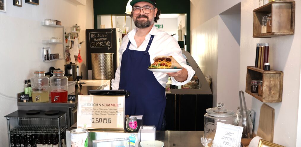 Ein Mann steht hinter der Theke und hält einen Teller mit einem Sandwich in der Hand.